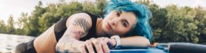Frau mit bleuen Haaren und Tattoos liegt auf einem Kajak im See